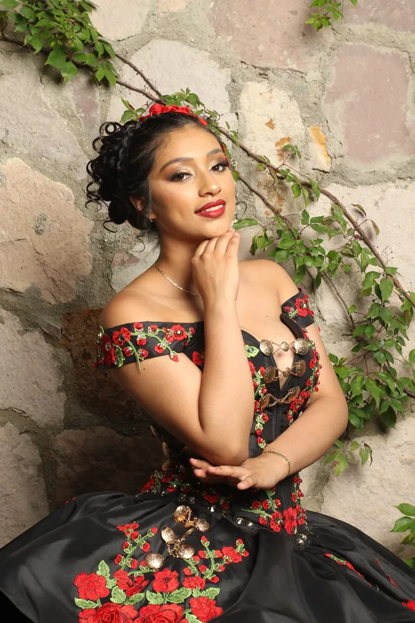 Antes: retrato de una quinceañera con vestido negro y flores rojas, recostada contra un muro de piedra con enredadera